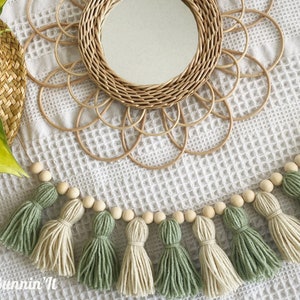 May include: A garland of wooden beads with green and cream tassels hangs on a white background. The garland is decorated with wooden beads and has a natural, bohemian style.
