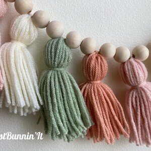 Op de afbeelding: Een guirlande van houten kralen en garenkwastjes in de kleuren roze, wit, groen en perzik. De guirlande is handgemaakt en heeft een "Just Bunnin' It" handtekening.