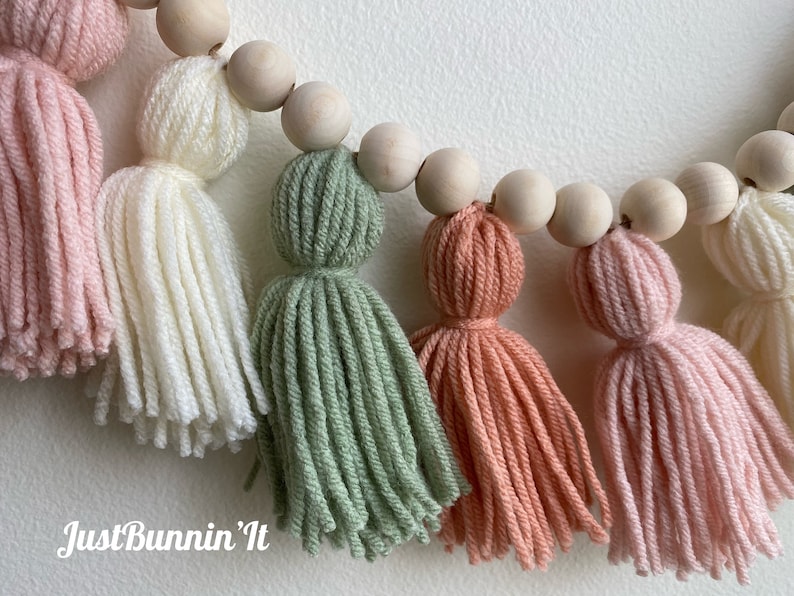 Op de afbeelding: Een guirlande van houten kralen en garenkwastjes in de kleuren roze, wit, groen en perzik. De guirlande is handgemaakt en heeft een "Just Bunnin' It" handtekening.