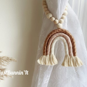 May include: A beige, white, and gold rainbow curtain tieback with a wooden bead garland. The rainbow is made of yarn and has a gold glitter accent.