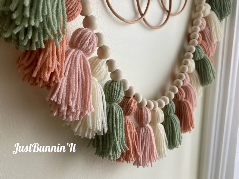Op de afbeelding: Een guirlande van houten kralen met kwastjes in tinten groen, roze en wit. De kwastjes zijn gemaakt van garen en zijn aan de kralen gebonden. De guirlande hangt aan een muur. De tekst "Just Bunnin' It" is zichtbaar in de linkerbenedenhoek van de afbeelding.