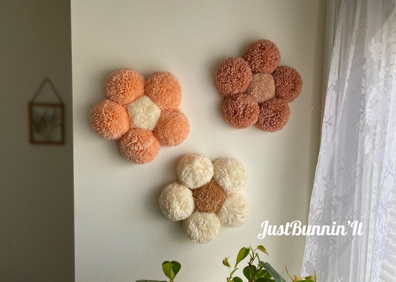 May include: Three handmade wall decor flowers made of yarn pom-poms. The flowers are in shades of peach, cream, and brown. The flowers are arranged on a white wall. The text "Just Bunnin' It" is visible in the bottom right corner of the image.