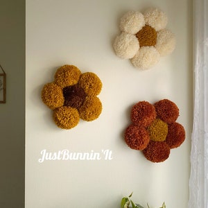 May include: Three yarn pom-pom flowers in different colors, mounted on a white wall. The flowers are in shades of yellow, orange, and white. The text "Just Bunnin' It" is written in white below the flowers.