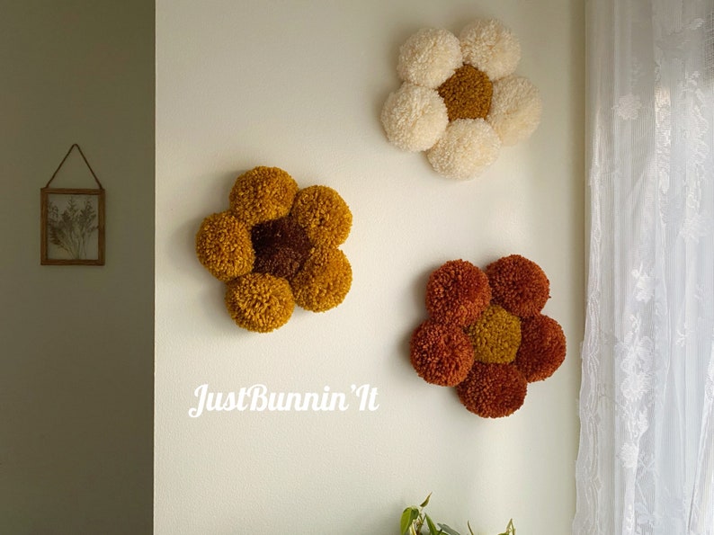 May include: Three yarn pom-pom flowers in different colors, mounted on a white wall. The flowers are in shades of yellow, orange, and white. The text "Just Bunnin' It" is written in white below the flowers.