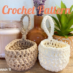 May include: Two crocheted hanging baskets with handles. One basket is beige and the other is white. The baskets are sitting on a wooden surface.