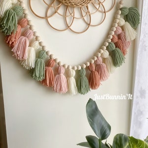 Op de afbeelding: Een guirlande van houten kralen met garenkwastjes in tinten groen, roze en wit. De guirlande hangt aan een muur met een geweven spiegel erachter. De tekst "Just Bunnin' It" is zichtbaar in de rechterbenedenhoek van de afbeelding.