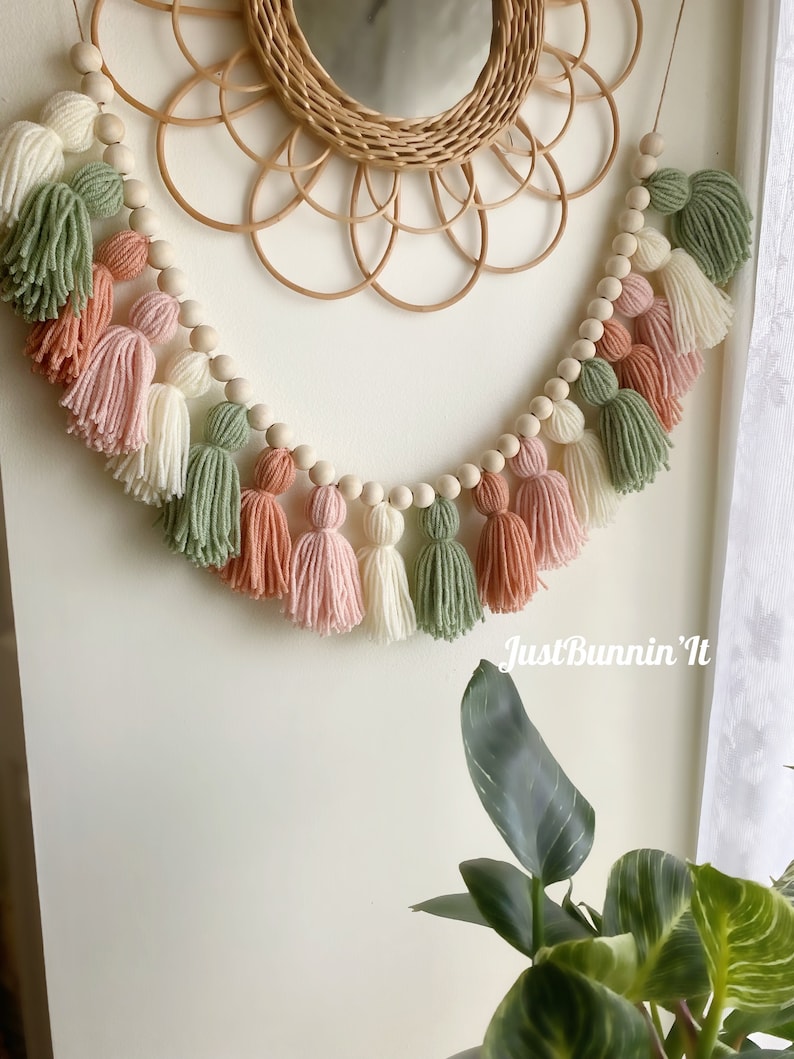 Op de afbeelding: Een guirlande van houten kralen met garenkwastjes in tinten groen, roze en wit. De guirlande hangt aan een muur met een geweven spiegel erachter. De tekst "Just Bunnin' It" is zichtbaar in de rechterbenedenhoek van de afbeelding.