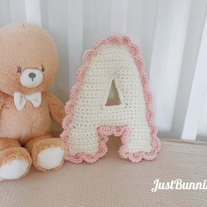 Könnte beinhalten: Ein Teddybär mit weißer Fliege sitzt neben einem gehäkelten Buchstaben "A" Kissen. Der Buchstabe ist cremefarben mit einem rosa, gewellten Rand. Der Text "JustBunnin'It" befindet sich unten rechts.