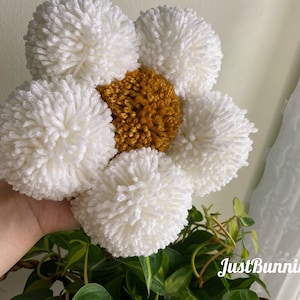 May include: A white yarn flower with a yellow center. The flower is made of pom-poms and is held in a hand.