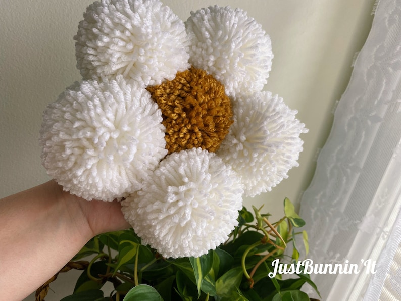 May include: A white yarn flower with a yellow center. The flower is made of pom-poms and is held in a hand.