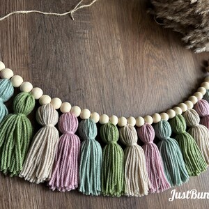 May include: A decorative tassel garland featuring a string of wooden beads and colorful yarn tassels. The tassels are in shades of green, pink, blue, and beige. The garland is designed for home decor.