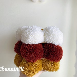 May include: A stack of three pom-poms in white, red, and yellow. The pom-poms are arranged in a pyramid shape.