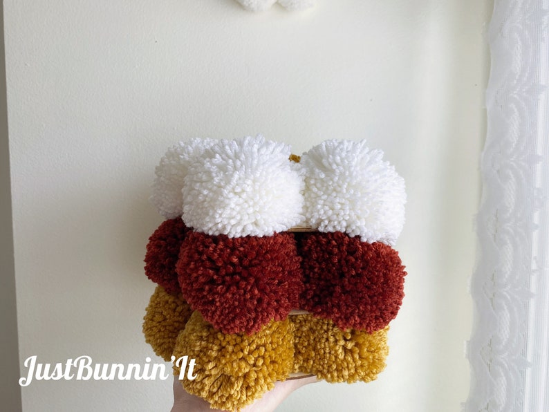 May include: A stack of three pom-poms in white, red, and yellow. The pom-poms are arranged in a pyramid shape.