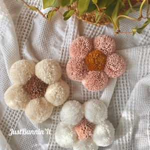 May include: Three handmade pom-pom flower wall decorations in pink, white, and cream colors. The flowers have a center circle of a different color. The text "Just Bunnin' It" is visible in the bottom left corner of the image.