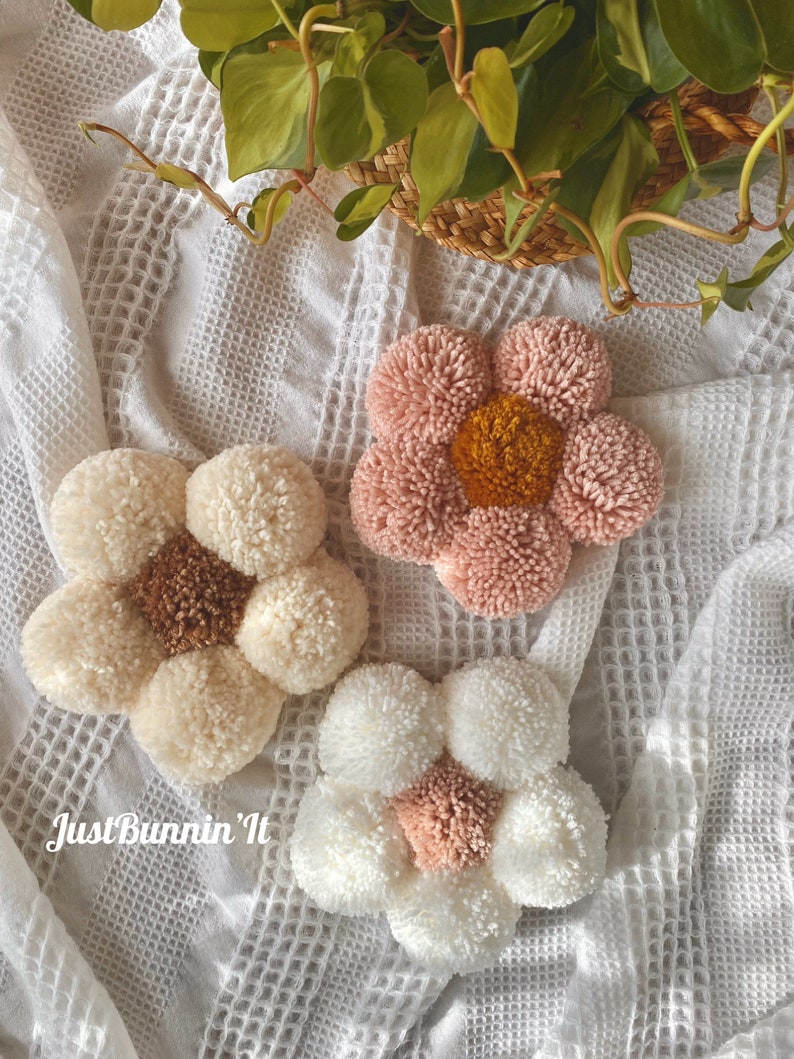 May include: Three handmade pom-pom flower wall decorations in pink, white, and cream colors. The flowers have a center circle of a different color. The text "Just Bunnin' It" is visible in the bottom left corner of the image.