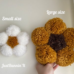 Op de afbeelding: Twee pompon-bloemen wanddecoraties, een klein en wit met een geel centrum, en een groot met een bruin centrum en gele bloemblaadjes. De tekst "Just Bunnin' It" staat onder de kleine bloem. De tekst "Large size" staat boven de grote bloem.