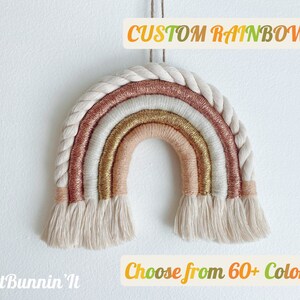 May include: A handmade rainbow wall hanging with a braided rope border and layers of gold, pink, and cream-colored yarn. The text "CUSTOM RAINBOW!" and "Choose from 60+ Colors" are displayed on the image. This decorative item is designed for home decor.