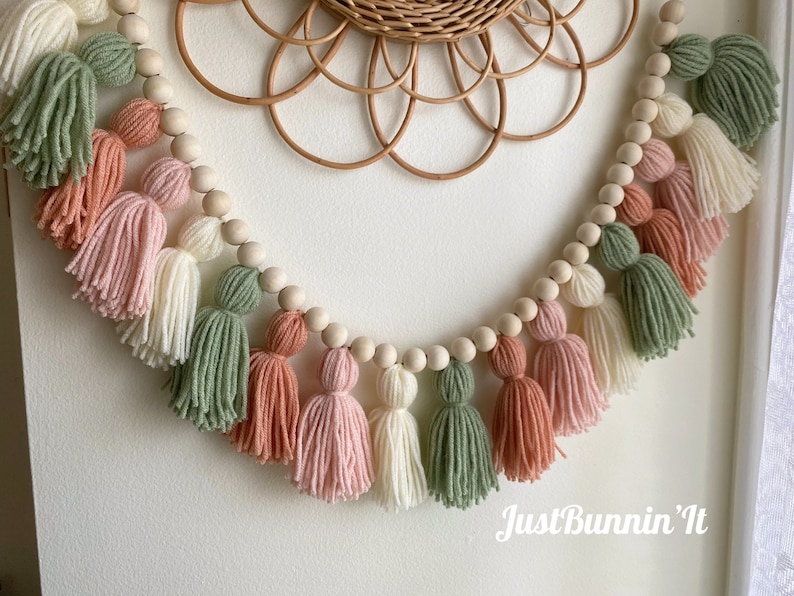 Op de afbeelding: Een guirlande van houten kralen met kleurrijke garenkwastjes in tinten groen, roze en wit. De guirlande hangt aan een witte muur met de tekst "Just Bunnin' It" onderaan.