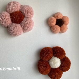 Op de afbeelding: Drie handgemaakte pomponbloemen in verschillende kleuren en maten. De grootste bloem is roze met een bruin centrum, de middelste bloem is perzikkleurig met een bruin centrum en de kleinste bloem is bruin met een wit centrum. De bloemen zijn bevestigd aan een witte muur.