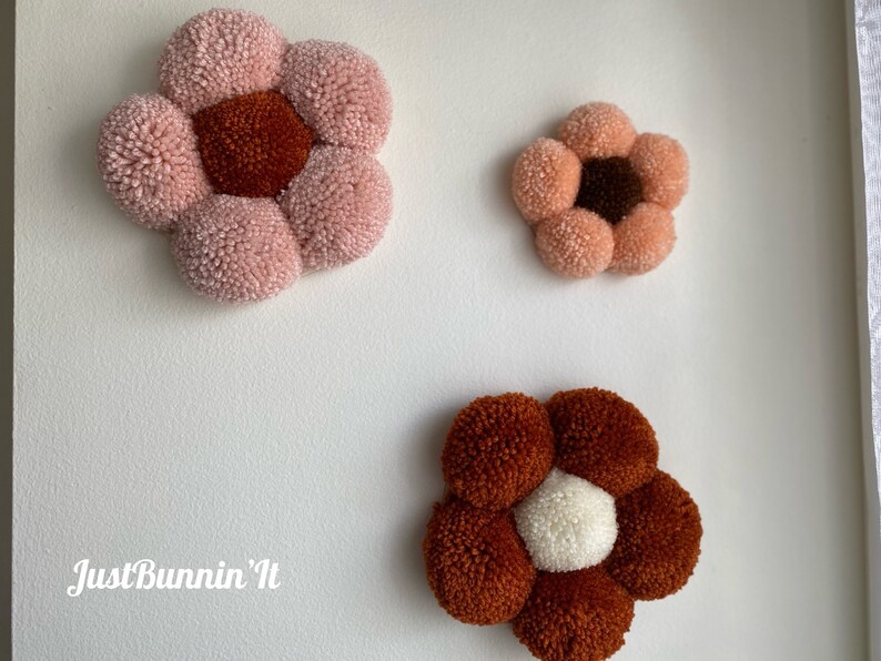Op de afbeelding: Drie handgemaakte pomponbloemen in verschillende kleuren en maten. De grootste bloem is roze met een bruin centrum, de middelste bloem is perzikkleurig met een bruin centrum en de kleinste bloem is bruin met een wit centrum. De bloemen zijn bevestigd aan een witte muur.