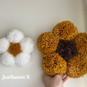 May include: Two handmade pom-pom flowers, one white with a yellow center and one yellow with a brown center. The flowers are made with yarn and are a popular craft project.