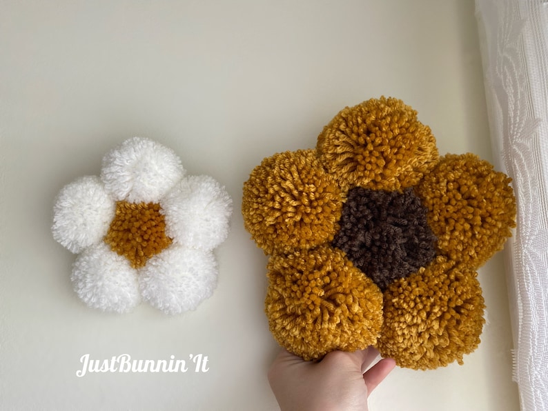 May include: Two handmade pom-pom flowers, one white with a yellow center and one yellow with a brown center. The flowers are made with yarn and are a popular craft project.