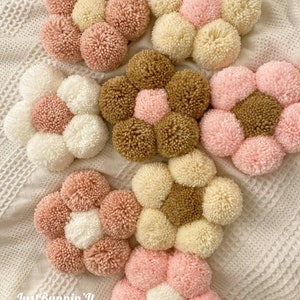 May include: Six handmade pom-pom flower wall decorations in pink, white, brown, and cream colors. The flowers are arranged in a circular pattern on a white crocheted surface.