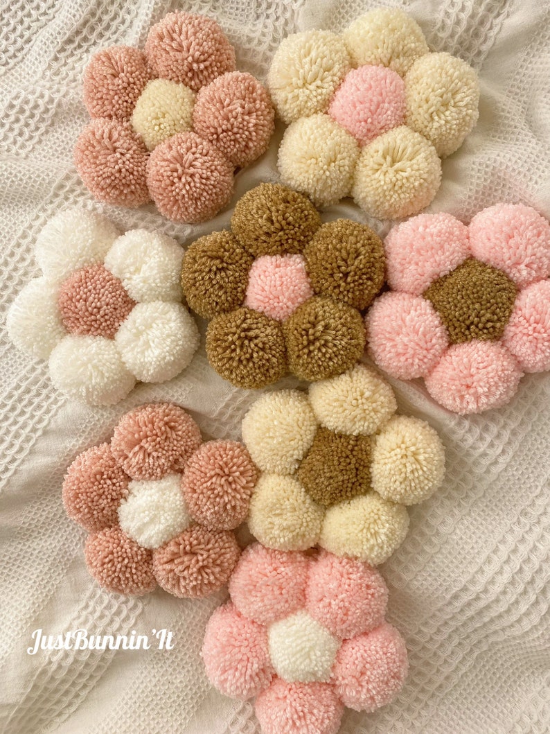 May include: Six handmade pom-pom flower wall decorations in pink, white, brown, and cream colors. The flowers are arranged in a circular pattern on a white crocheted surface.