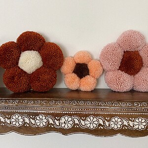 Op de afbeelding: Drie kleurrijke bloemvormige pomponkussens in tinten roze, oranje en bruin, getoond op een houten plank met een decoratieve witte rand.
