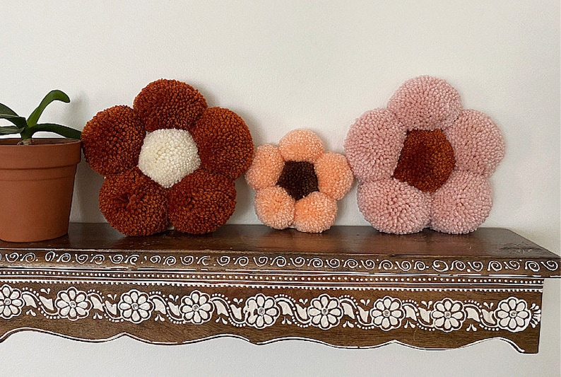 Op de afbeelding: Drie kleurrijke bloemvormige pomponkussens in tinten roze, oranje en bruin, getoond op een houten plank met een decoratieve witte rand.