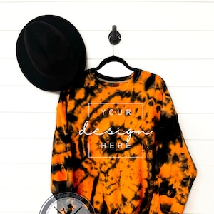 Puede incluir: Sudadera naranja y negra tie-dye con el texto "YOUR design HERE" en la parte delantera. La sudadera está colgada de una percha sobre un fondo de madera blanca.