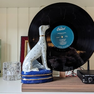 May include: A white and black spotted ceramic Dalmatian statue with a gold collar sits on a blue cushion with gold trim. A vintage vinyl record with a blue label is displayed behind the statue. A small black audio amplifier is on the shelf.