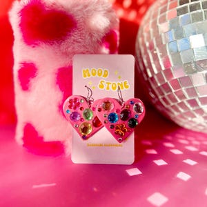 May include: A pair of pink heart-shaped earrings attached to a display card labeled MOOD STONE HANDMADE ACCESSORIES. The earrings are decorated with various colorful, faceted rhinestones in shapes including circles and flowers, featuring silver hooks.