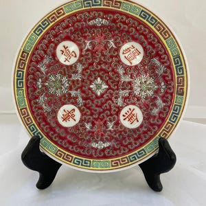 May include: A round ceramic decorative plate featuring a deep red background with intricate white floral scrollwork and four white circular medallions containing red Chinese characters. The rim has a colorful geometric border pattern.