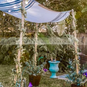 May include: Outdoor canopy with a white and blue striped fabric roof, supported by wooden poles. The canopy is decorated with string lights and greenery. Potted plants and a turquoise planter are visible in the foreground, creating a garden setting.