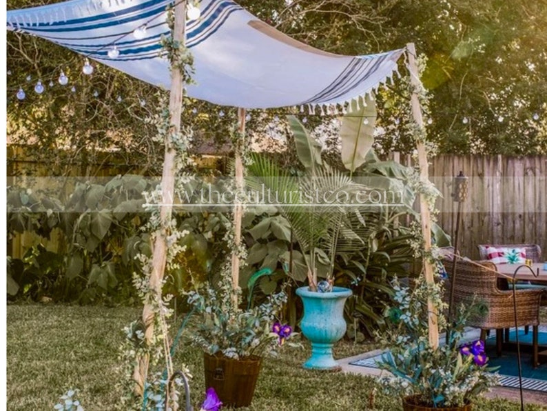 May include: Outdoor canopy with a white and blue striped fabric roof, supported by wooden poles. The canopy is decorated with string lights and greenery. Potted plants and a turquoise planter are visible in the foreground, creating a garden setting.