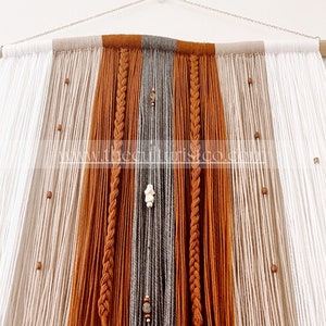 May include: A large, handmade wall hanging featuring a combination of white, tan, gray, and rust-colored yarn. The design includes braided sections and wooden beads, suspended from a natural wood dowel and chain.