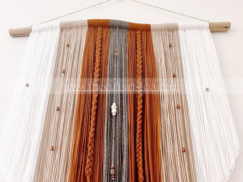May include: A large, handmade wall hanging featuring a combination of white, tan, gray, and rust-colored yarn. The design includes braided sections and wooden beads, suspended from a natural wood dowel and chain.