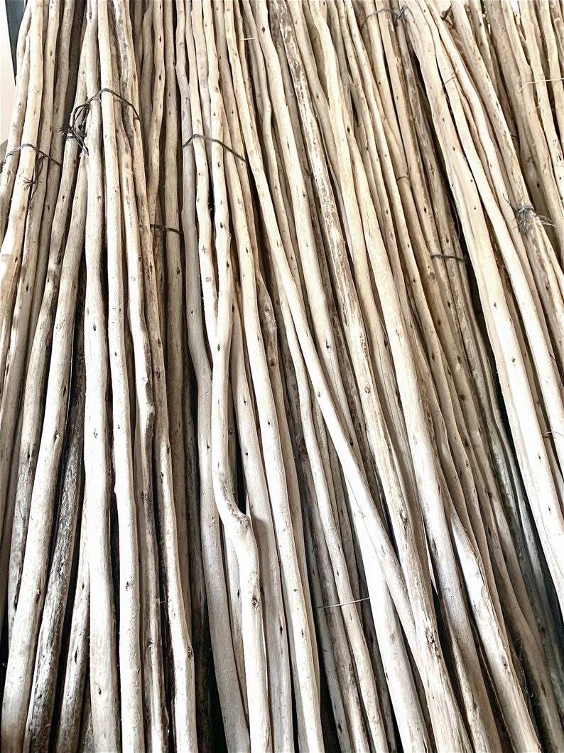 May include: A bundle of natural wooden poles, varying in thickness and length. The poles are light brown with some darker areas and knots, tied together with dark twine. The arrangement suggests a rustic or natural decor theme.