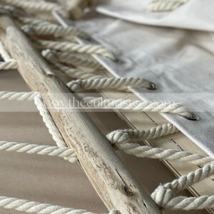 May include: Close-up of a handcrafted macrame piece featuring a natural wooden dowel and thick, off-white cotton rope. The design includes a woven pattern and a canvas fabric with metal grommets. The overall aesthetic is rustic and bohemian.
