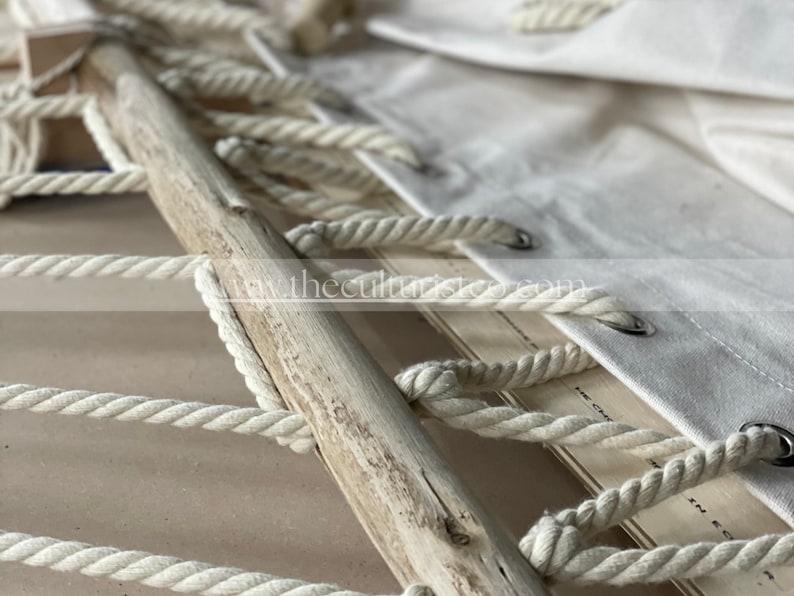 May include: Close-up of a handcrafted macrame piece featuring a natural wooden dowel and thick, off-white cotton rope. The design includes a woven pattern and a canvas fabric with metal grommets. The overall aesthetic is rustic and bohemian.