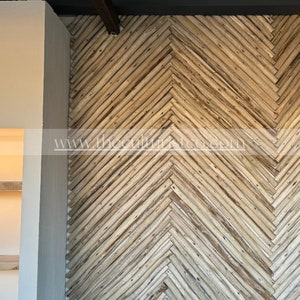 May include: A wall with a herringbone pattern made of light brown wooden planks. The planks are arranged diagonally, creating a textured surface. The wall is part of an interior design, with a neutral-colored wall and a dark gray border.