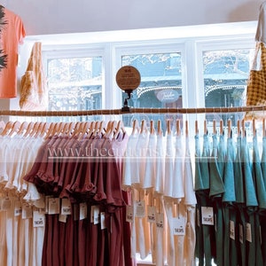 May include: Clothing display featuring a variety of t-shirts and dresses in shades of white, burgundy, and teal. Some shirts have tie-dye patterns. The items are hanging on wooden hangers from a horizontal rod. The text "THE DAYS" is visible on the tags.