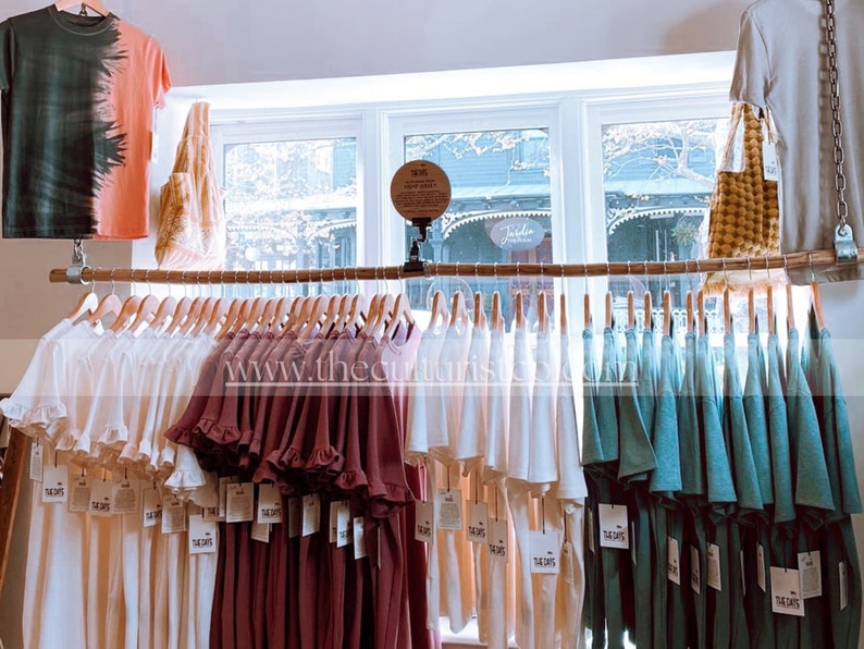 May include: Clothing display featuring a variety of t-shirts and dresses in shades of white, burgundy, and teal. Some shirts have tie-dye patterns. The items are hanging on wooden hangers from a horizontal rod. The text "THE DAYS" is visible on the tags.