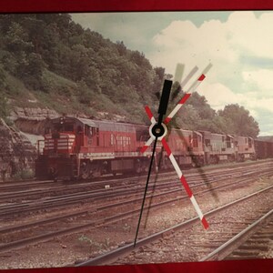 May include: A red and white train with a clock face superimposed on the image. The train is on a set of railroad tracks with a green hillside in the background.