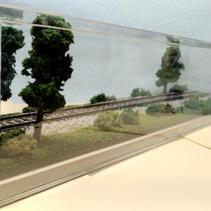 May include: A miniature model railroad scene in a clear plastic display case. The scene features a small train track with green foliage and trees on either side.