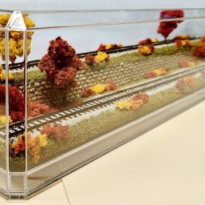 May include: A miniature model railway scene in a clear acrylic display case. The scene features a stone wall, two sets of railway tracks, and a variety of autumn foliage in shades of red, orange, and yellow.