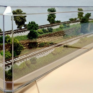 May include: A miniature model railroad scene with two sets of tracks, a stone wall, and green foliage, all enclosed in a clear plastic display case.