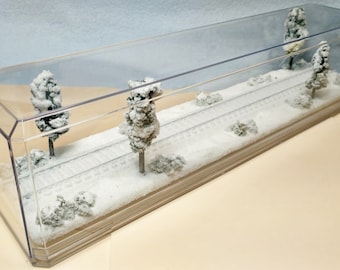 Op de afbeelding: Een miniatuur winterlandschap in een heldere acryl display case. De scène toont een besneeuwd landschap met modelbomen, struiken en treinrails. De bomen en struiken zijn bedekt met witte sneeuw.
