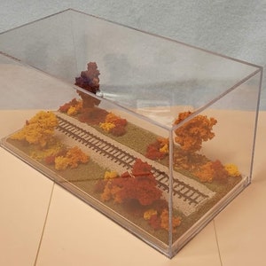 May include: A miniature train track scene with fall foliage in a clear acrylic display case. The track is surrounded by small trees with red, orange, and yellow leaves. The base of the display is covered in brown sand.